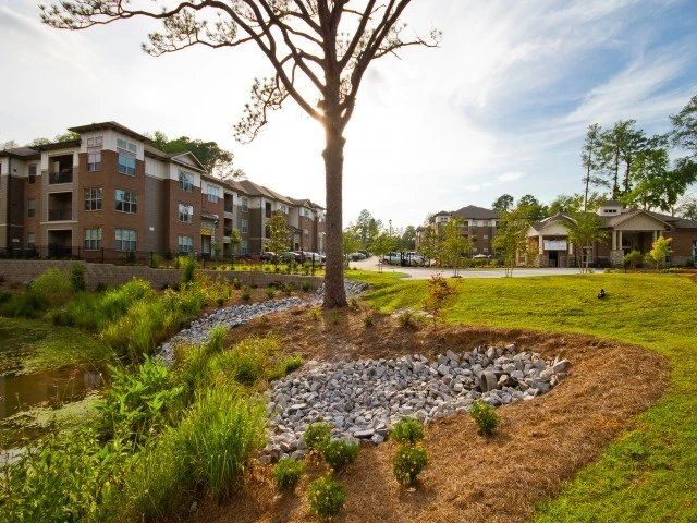 Apartment buildings beside a landscaped pond with rocks, grass, and trees on a sunny day  at Arcadia's Edge Apartments in Columbia, SC