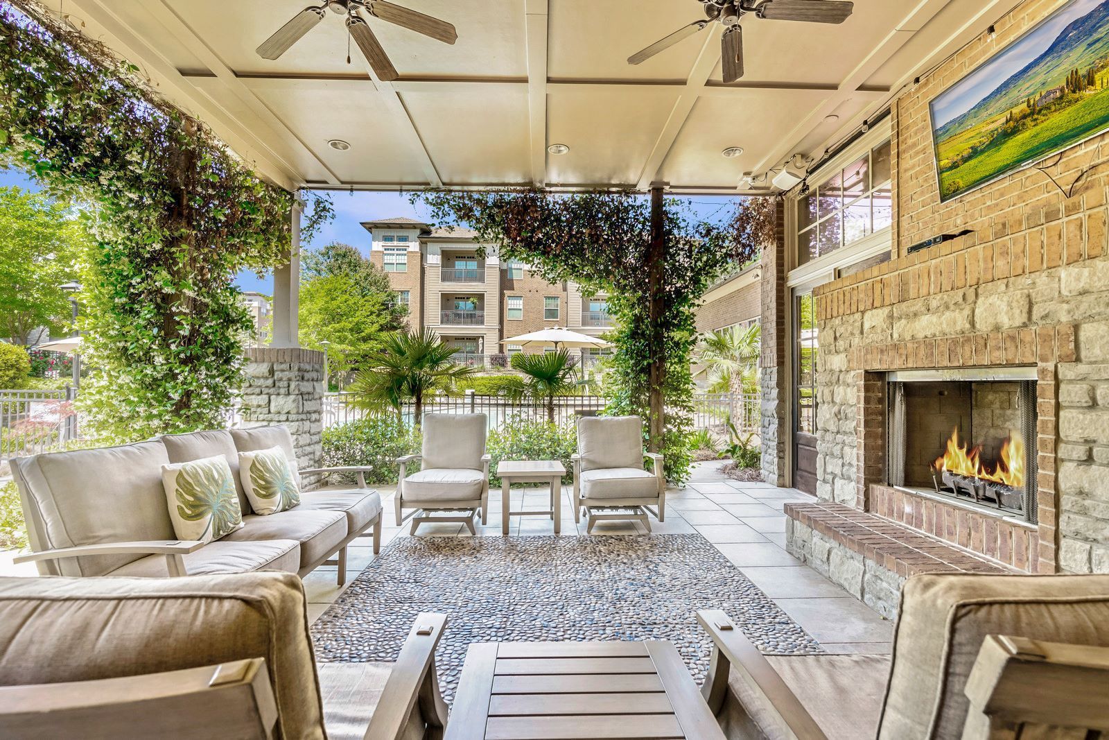 Covered patio with wicker seating, patterned rug, and stone fireplace overlooking a landscaped garden  at Arcadia's Edge Apartments in Columbia, SC