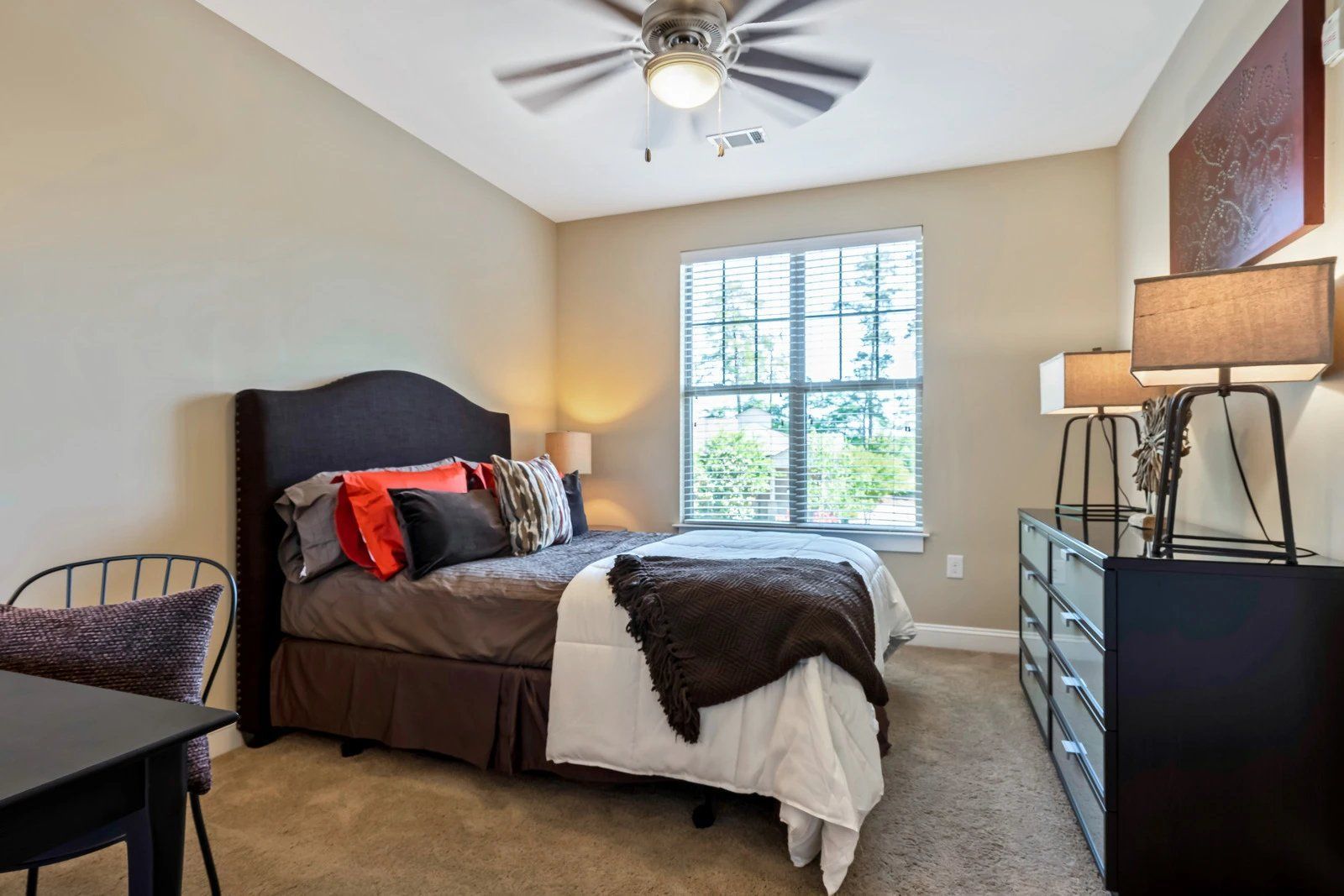 Bedroom with dark bed, red accent pillows, dresser, chair, lamp, and window with blinds  at Arcadia's Edge Apartments in Columbia, SC