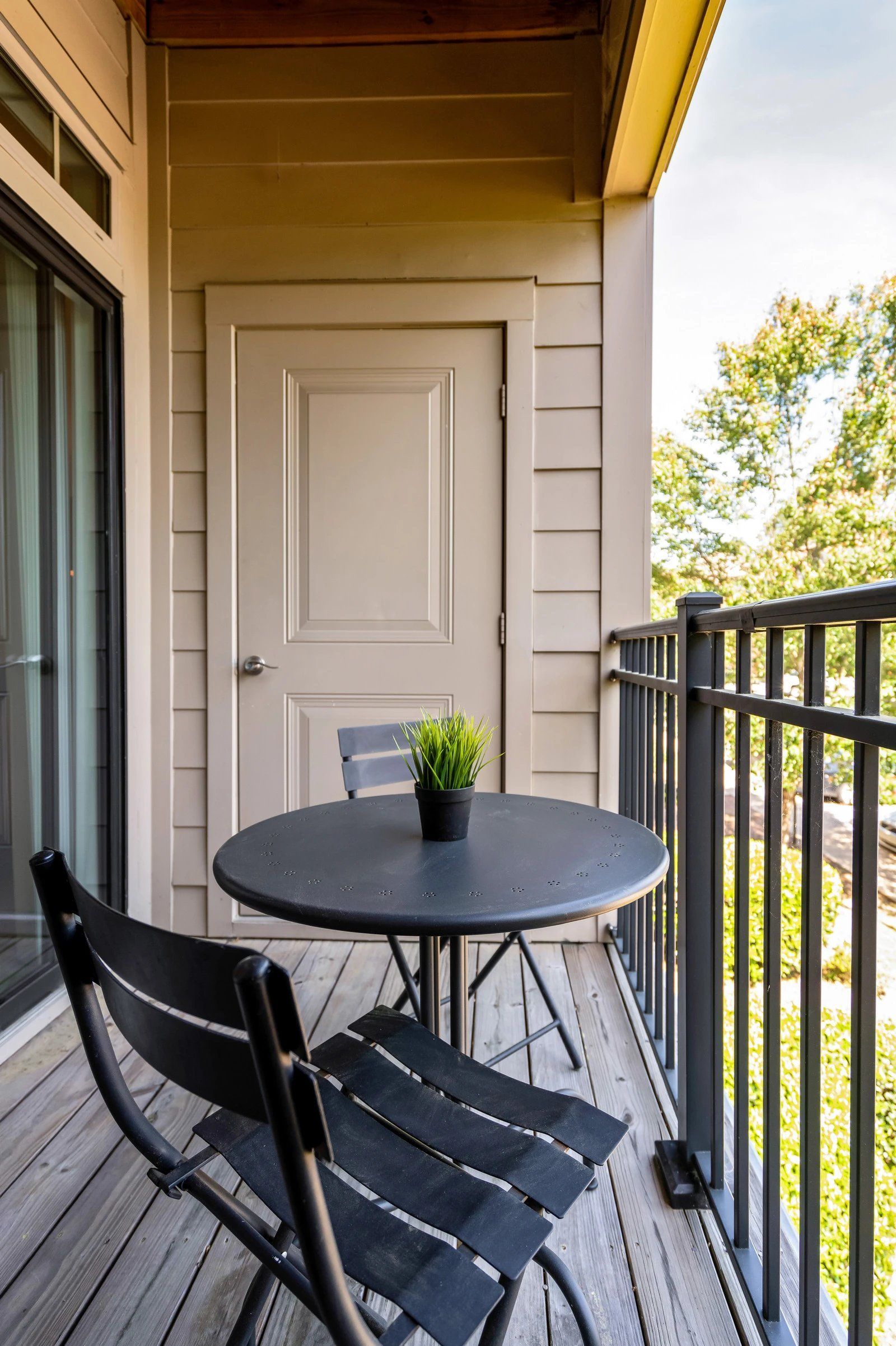 Small balcony with a round black table, chairs, and a potted plant beside a white door  at Arcadia's Edge Apartments in Columbia, SC