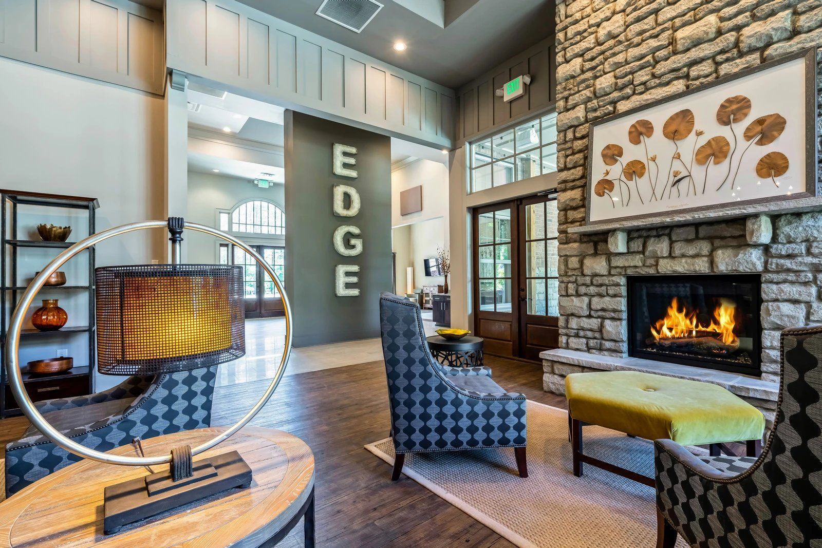 Modern lobby lounge with stone fireplace, blue chairs, wicker lamp, and large “EDGE” wall sign  at Arcadia's Edge Apartments in Columbia, SC