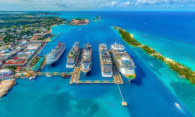 Cruise ships docked in a turquoise harbor, with a city and palm tree-lined island in the background.