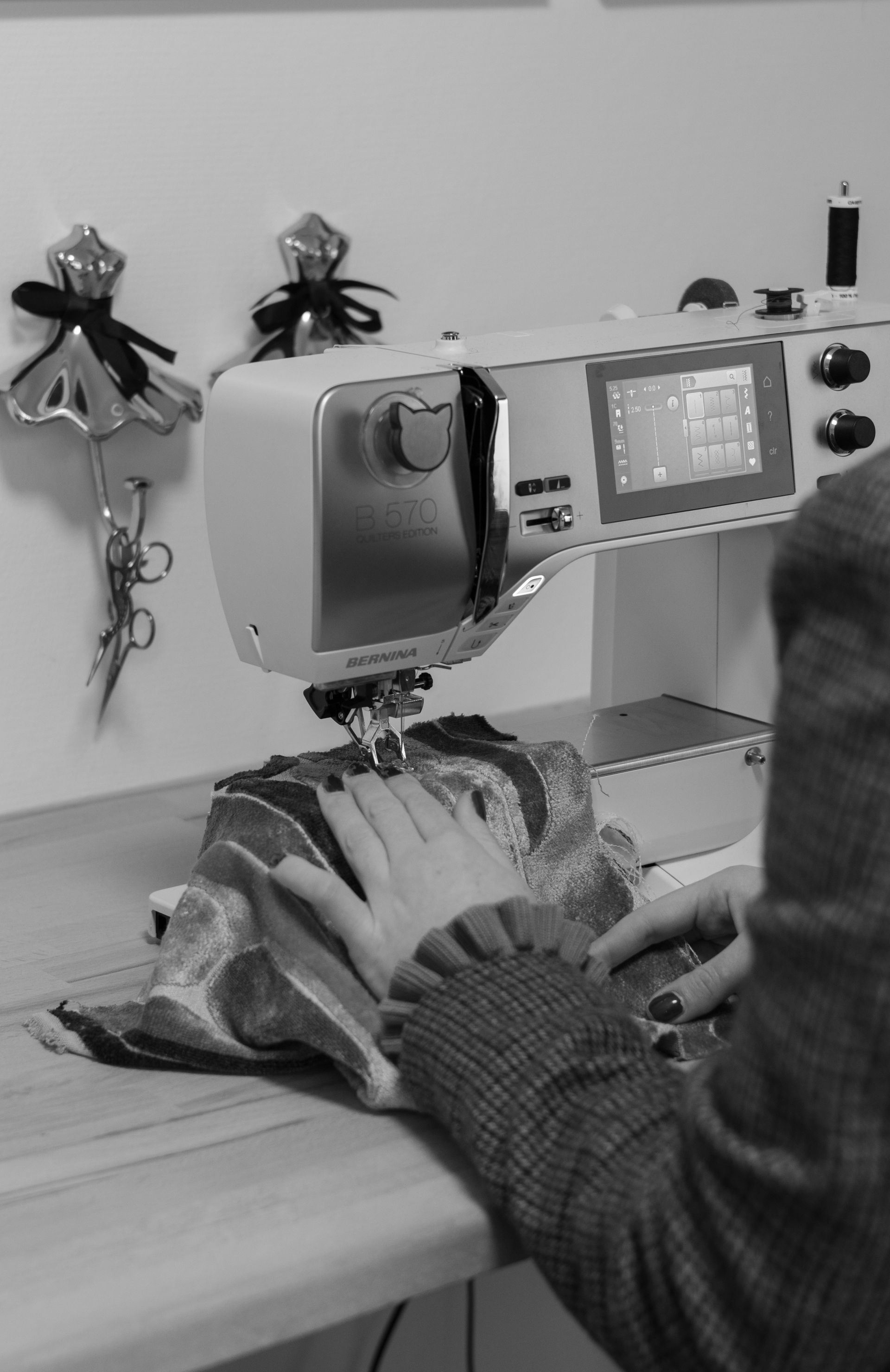 Een persoon naait aan een naaimachine naast twee kleine wanddecoraties in een werkplaatsomgeving.