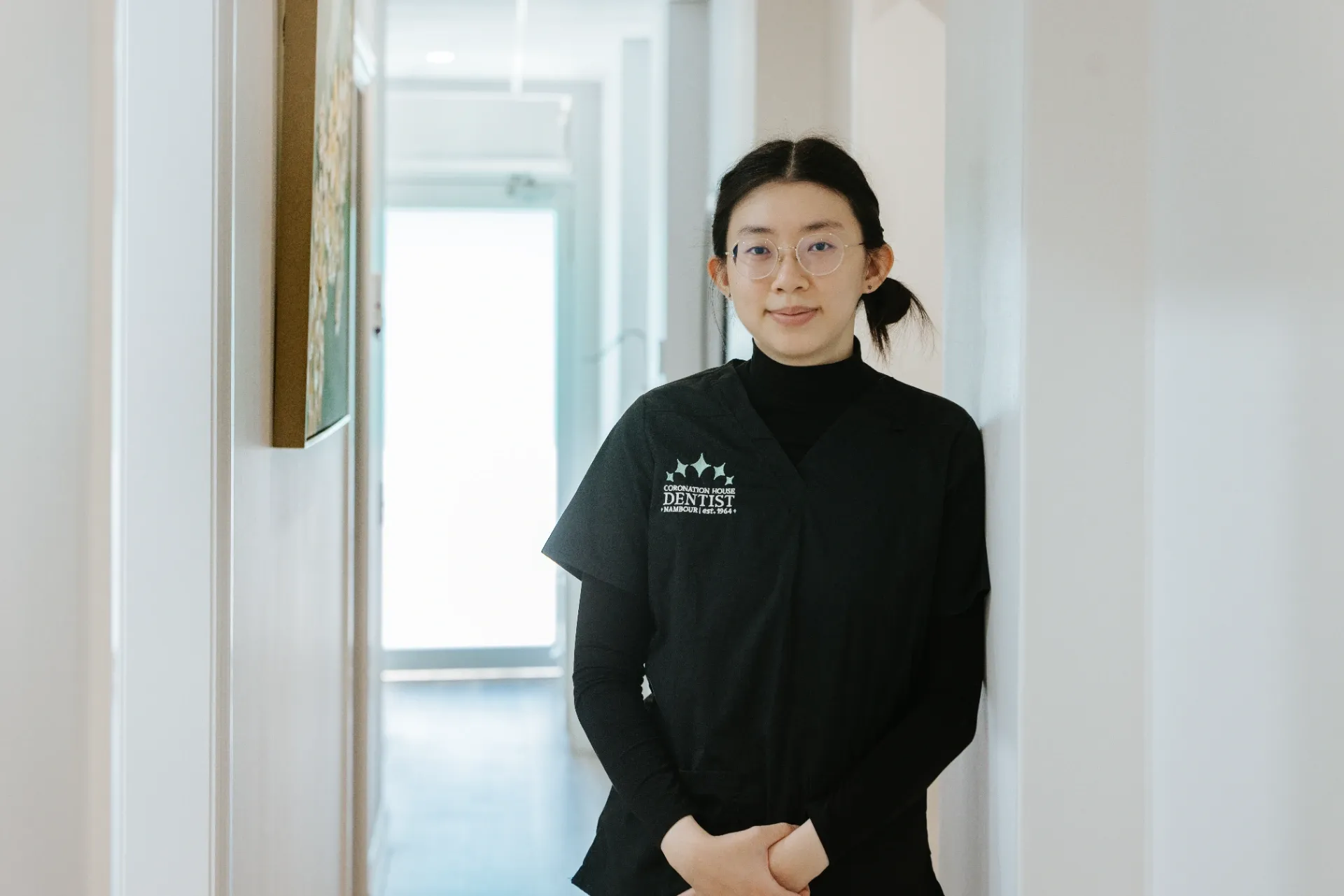 Person in a black shirt standing in a bright hallway, hands clasped, facing the camera. — Coronation House Dentist in Nambour, QLD