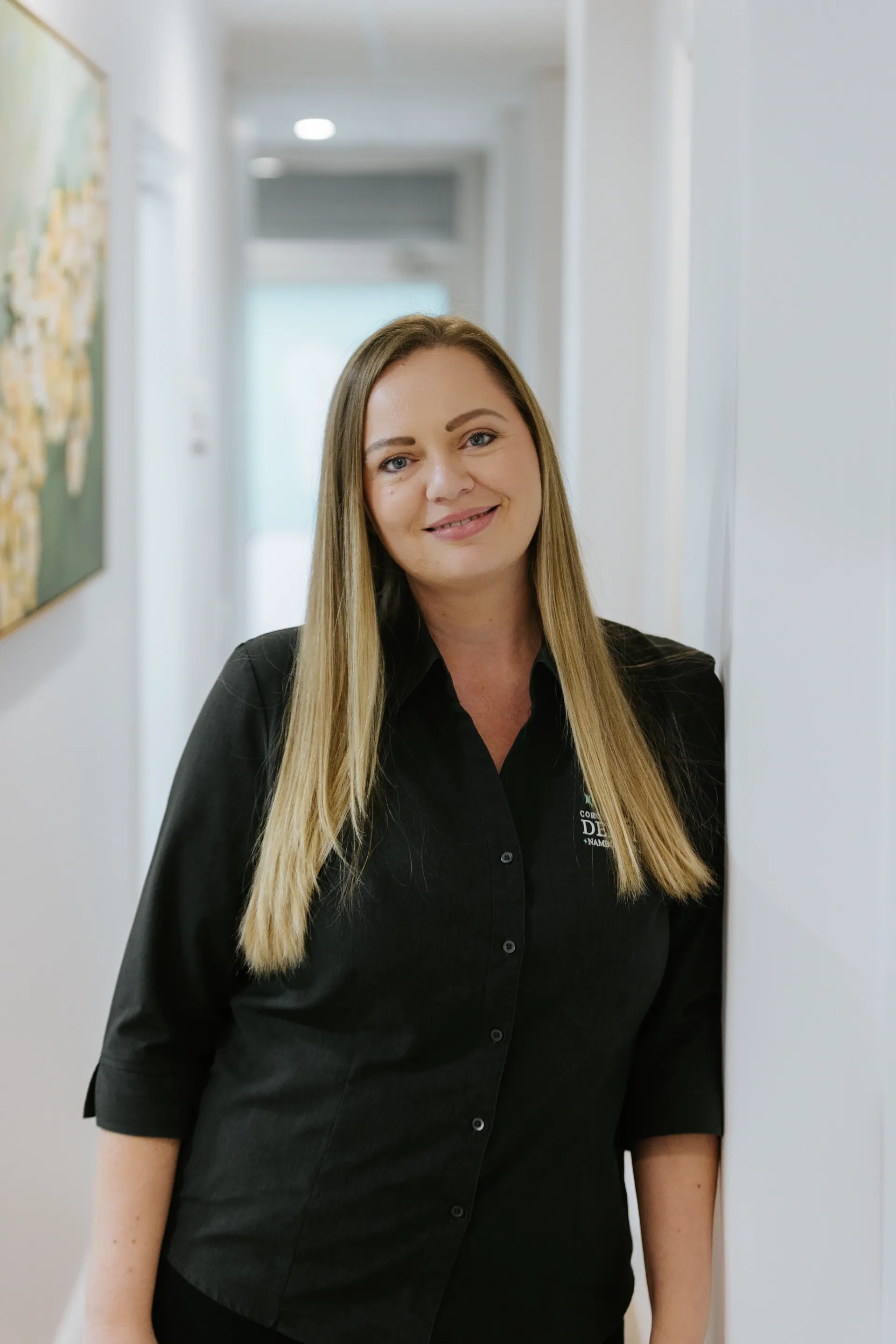 Woman in a black shirt standing in a bright hallway, smiling at the camera.