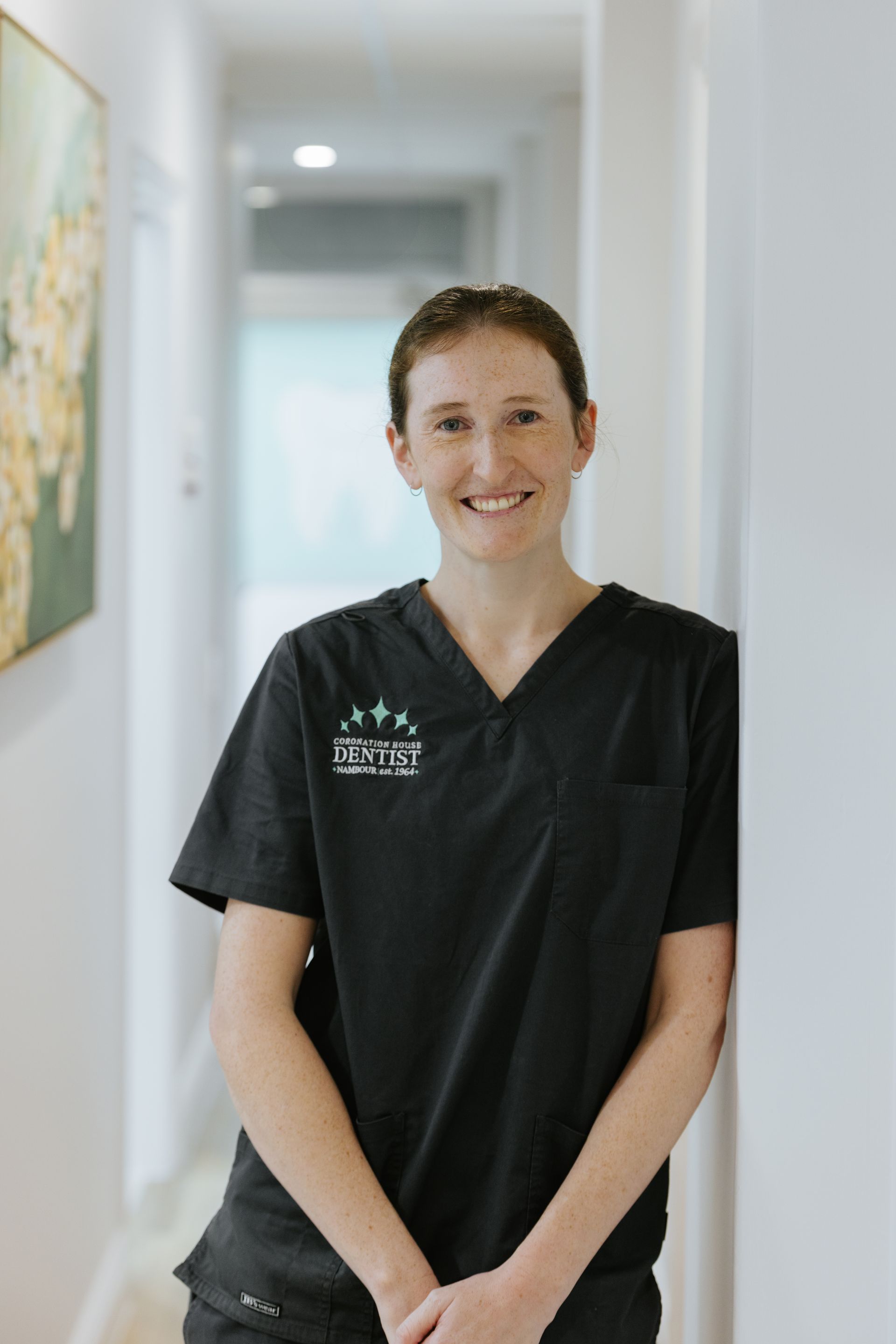 Person in black scrubs standing in a bright hallway, smiling with hands clasped.