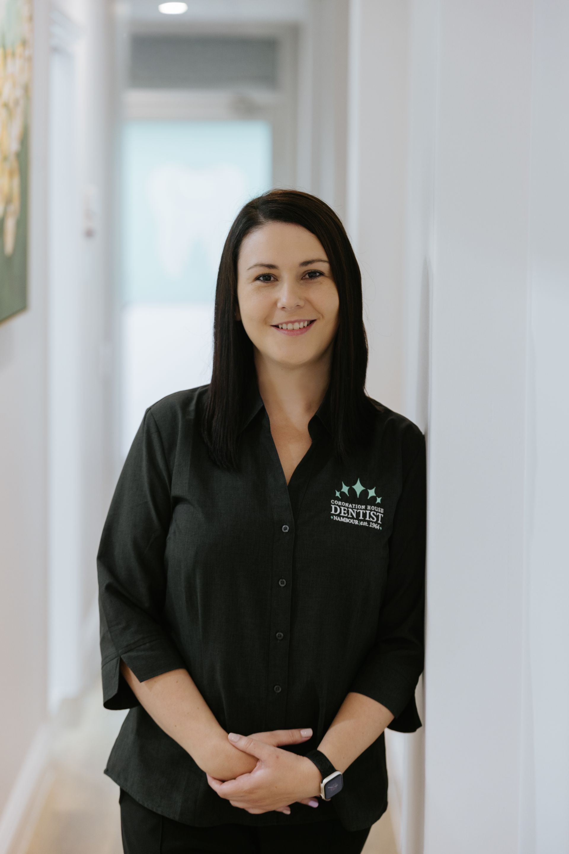Woman in a black shirt standing in a bright hallway, smiling at the camera.