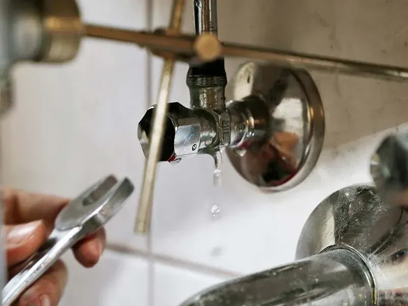 A hand holding a wrench near a leaky pipe shut-off valve under a sink.