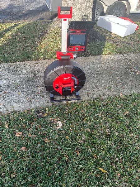 A red and black Milwaukee sewer inspection camera system stands on a sidewalk next to grass.