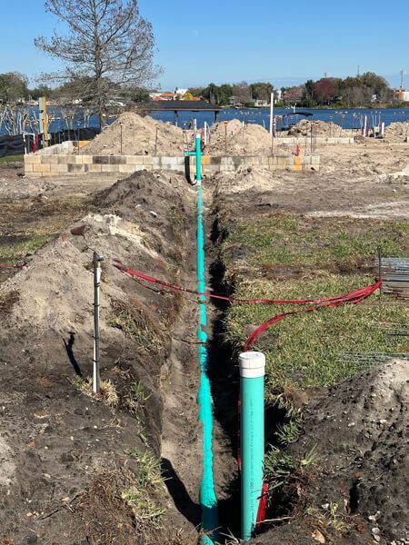 A long trench with green PVC pipes runs through a construction site toward a foundation near a waterfront.