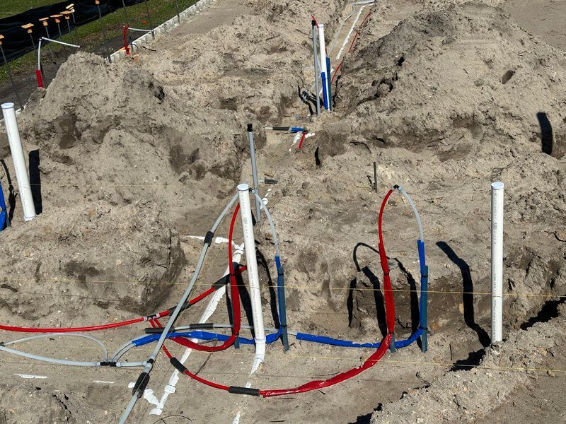 Construction site showing red and blue PEX plumbing pipes emerging from sandy ground around white vertical conduits.