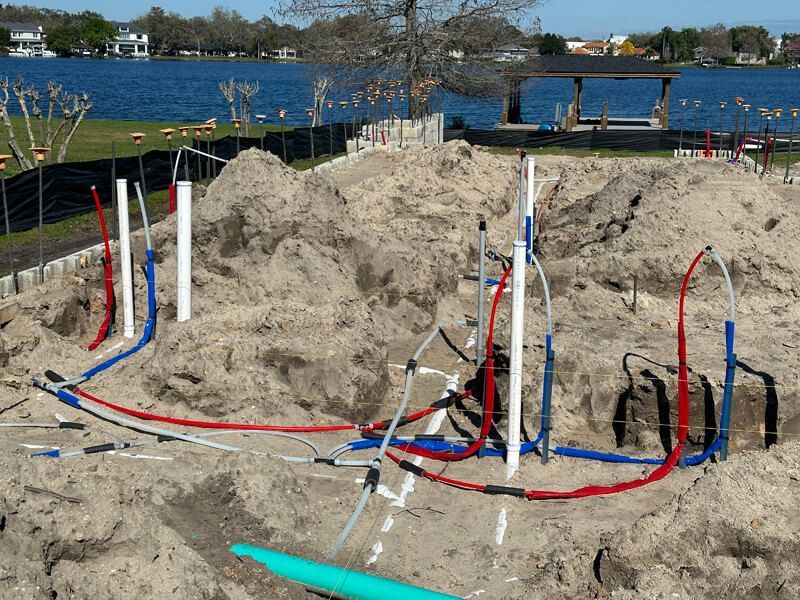 Construction site near a lake featuring exposed white pipes and colorful red and blue cables routed through sandy ground.