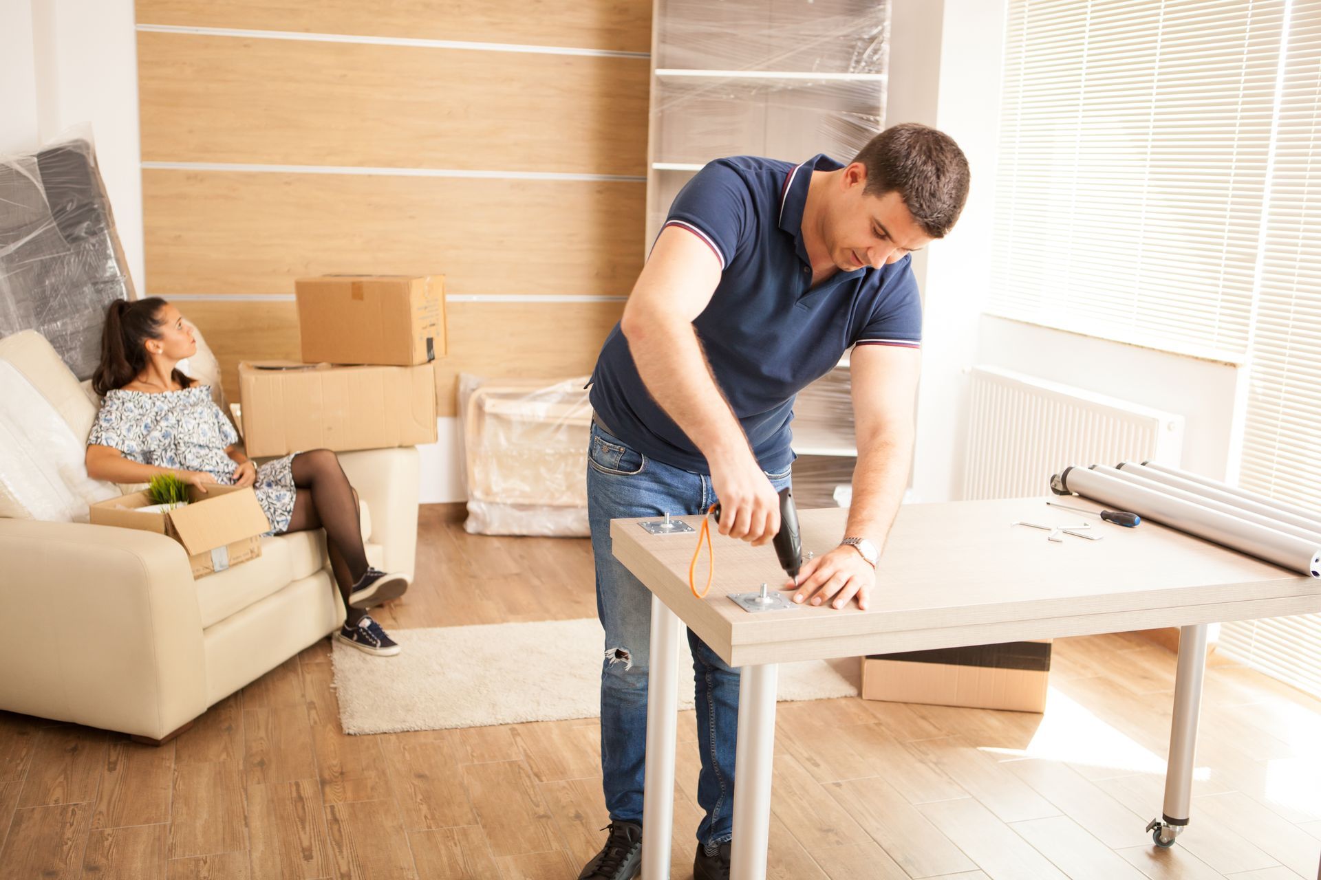 Man unpacking a table in a bright room while another person sits on a sofa among moving boxes
