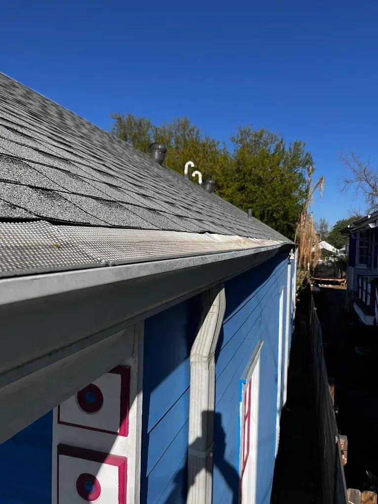 Hand clearing wet leaves and debris from a clogged roof gutter beside gray shingles