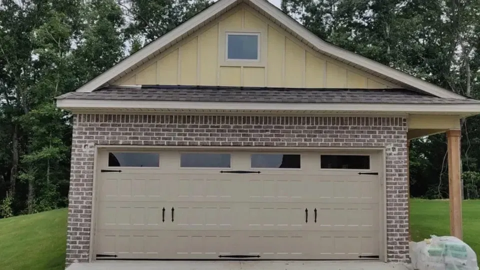 Tan garage door with brick and yellow siding, small window, and brown roof.
