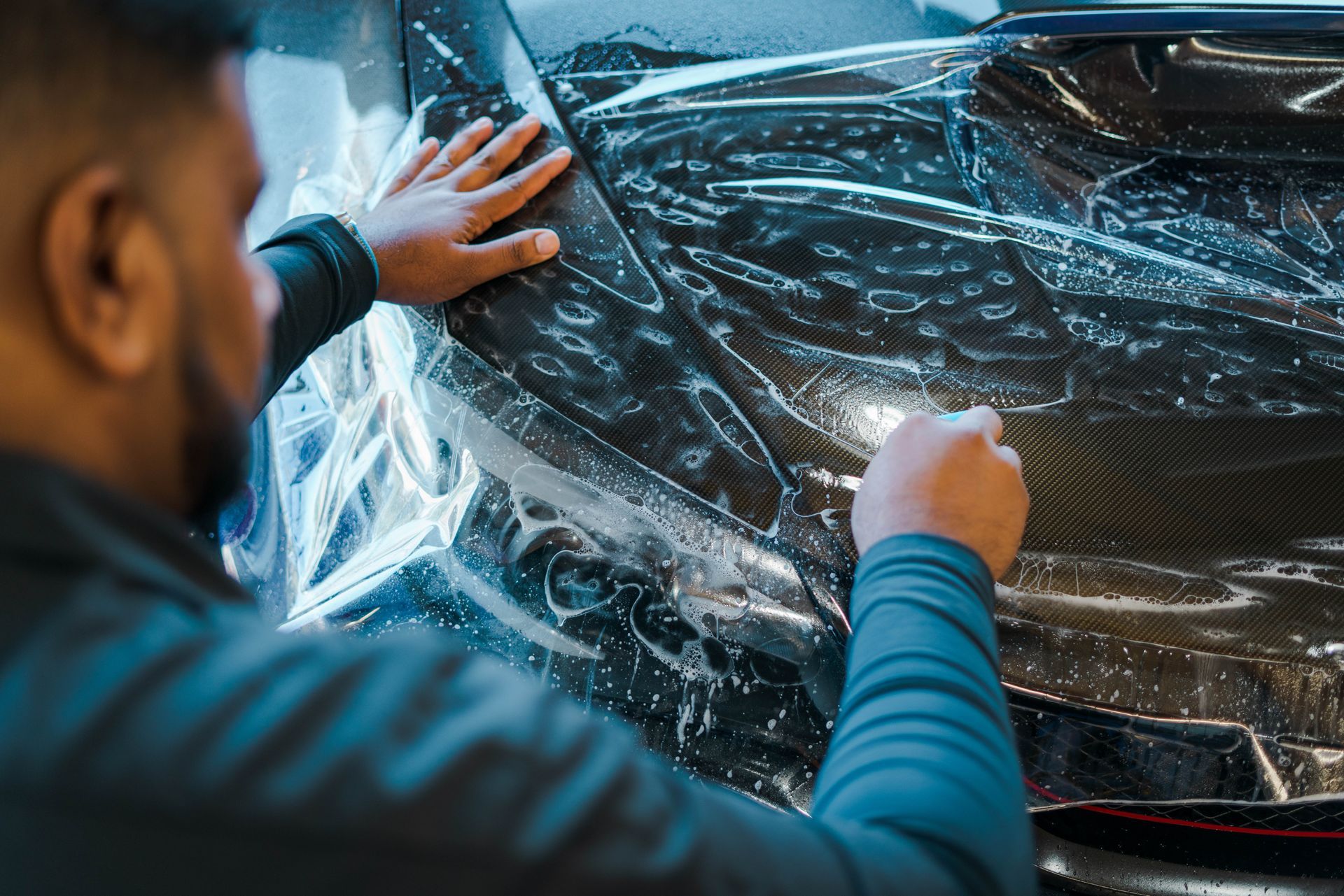 Man applying clear protective film to a black car hood, indoors.