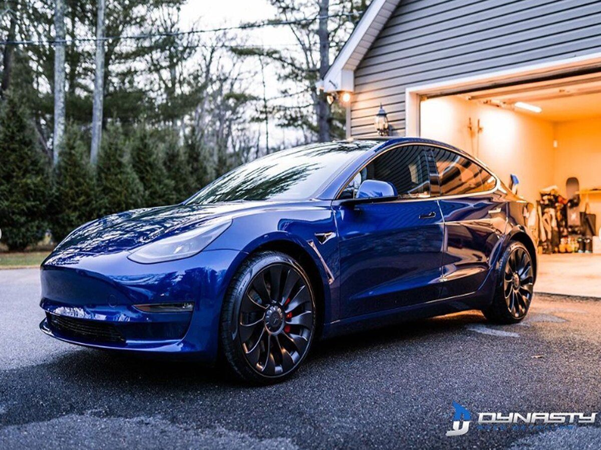 A blue tesla model 3 is parked in front of a garage.