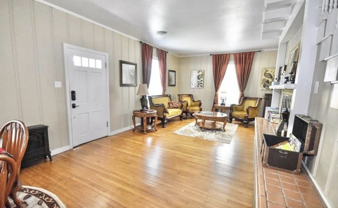 Bright living room with hardwood floors, white walls, yellow sofa, chairs, fireplace, and tall windows with curtains