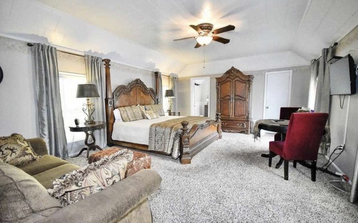 Bright bedroom with large bed, gray sofa, red chair, wardrobe, and ceiling fan on a white carpet.