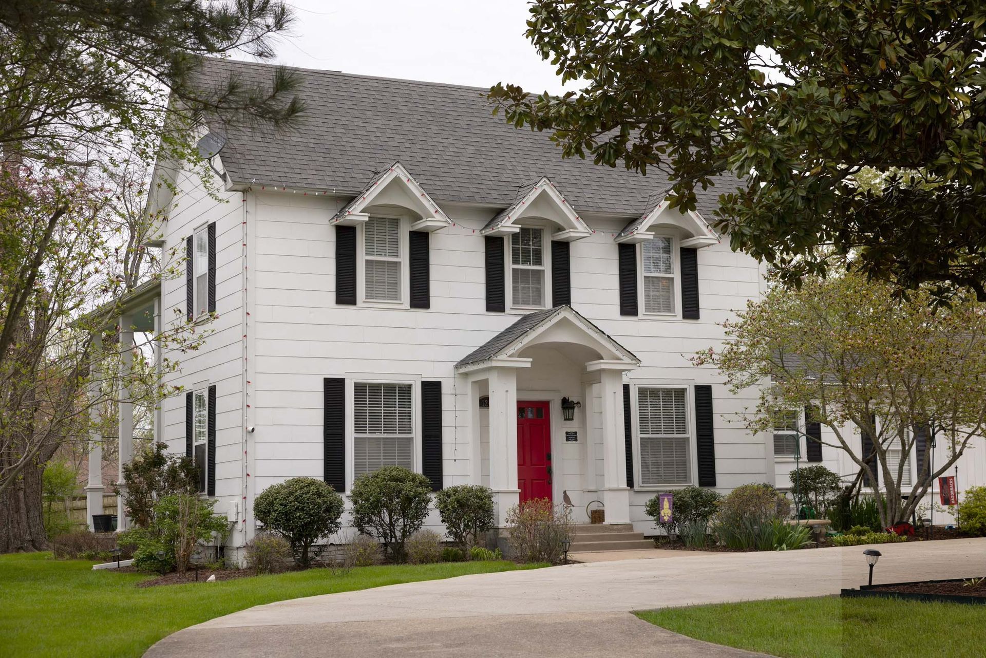 Two-story white house with black shutters and a red front door, framed by trees and a curved driveway