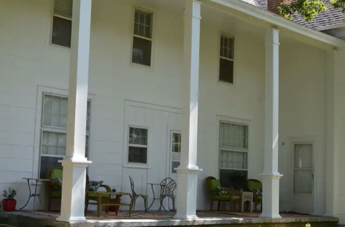 White house porch with tall columns, potted plants, and chairs along the front.