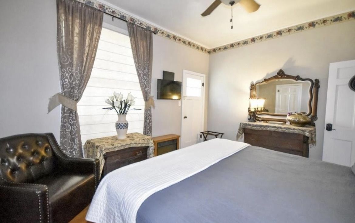 Bright bedroom with a bed, leather chair, dresser, mirror, and curtained window