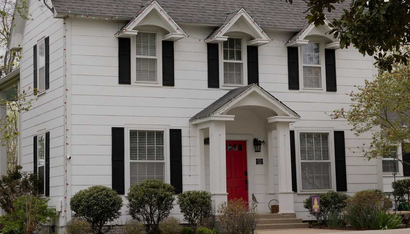 White two-story house with black shutters and a bright red front door, surrounded by shrubs and trees