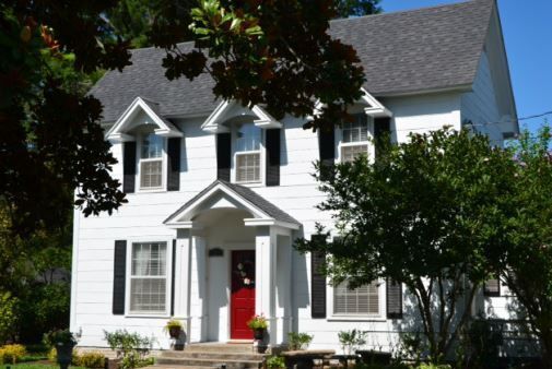 White two-story house with red front door, black shutters, and trees in front
