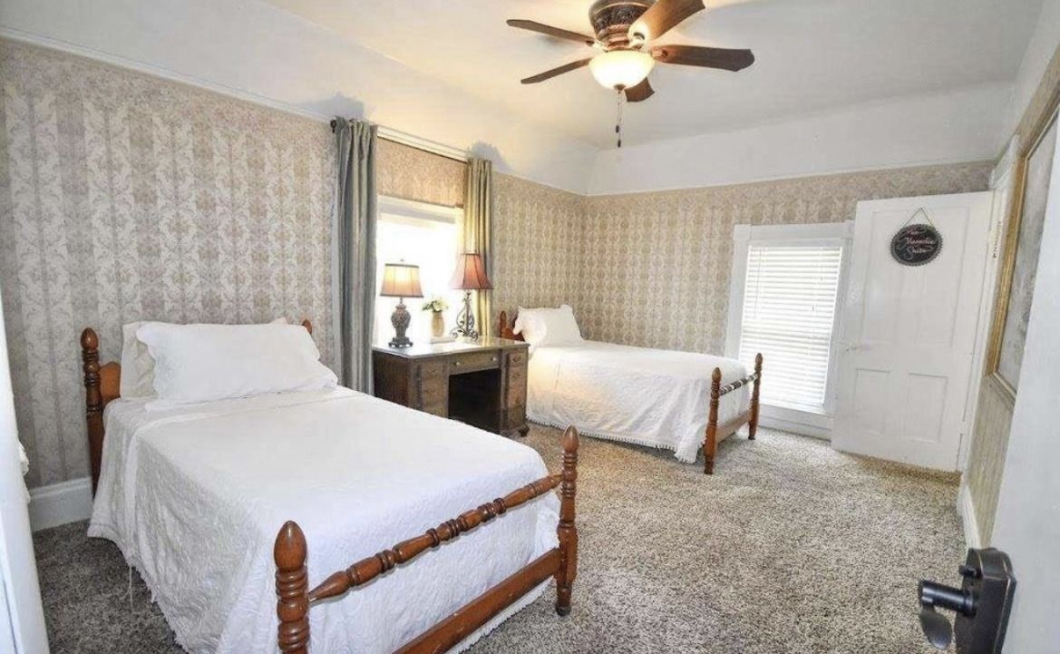 Bright bedroom with two white twin beds, patterned curtains, ceiling fan, and carpeted floor