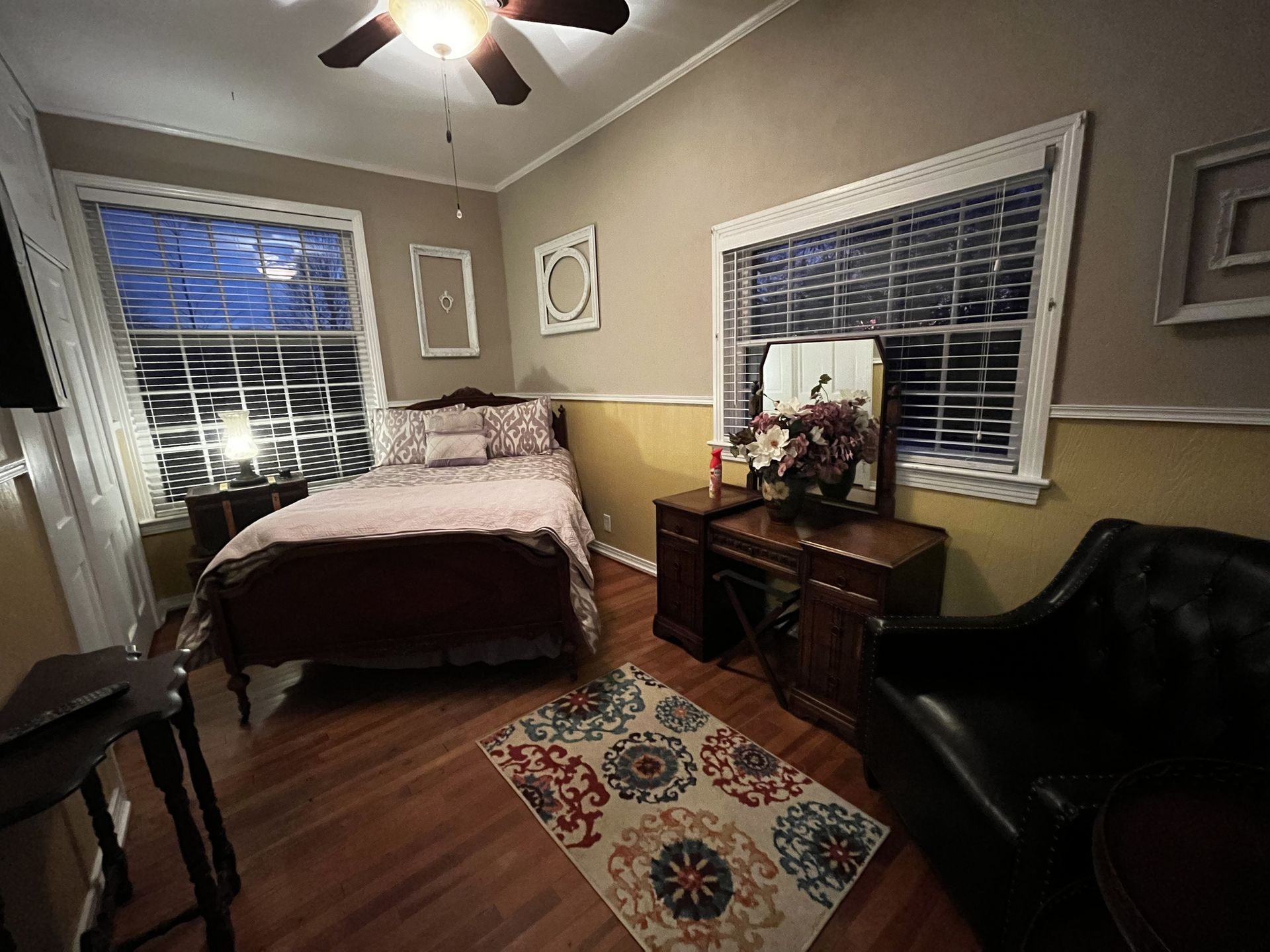 Bedroom with bed, two windows, dresser with mirror, black chair, and patterned rug on wood floor.