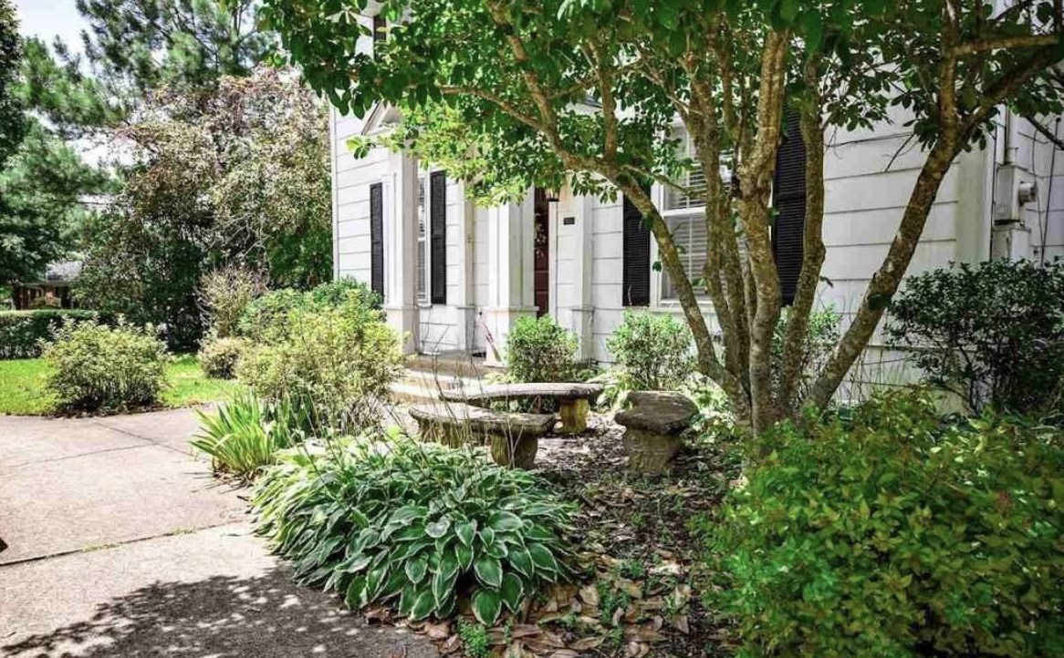 Garden path beside a white house with shrubs, trees, and a stone bench