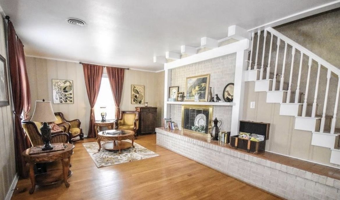 Cozy living room with hardwood floors, white staircase, fireplace mantel, and red curtains.