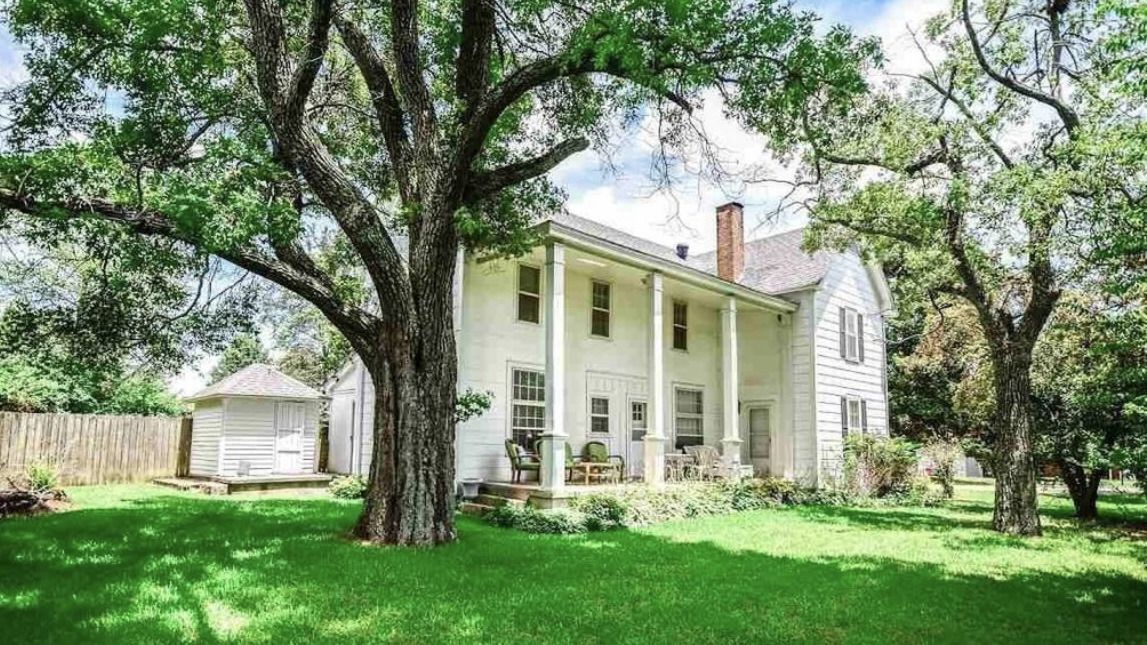 White two-story house with a front porch and large trees in a green yard