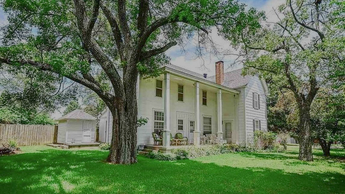 White farmhouse with large trees and green lawn under a bright sky