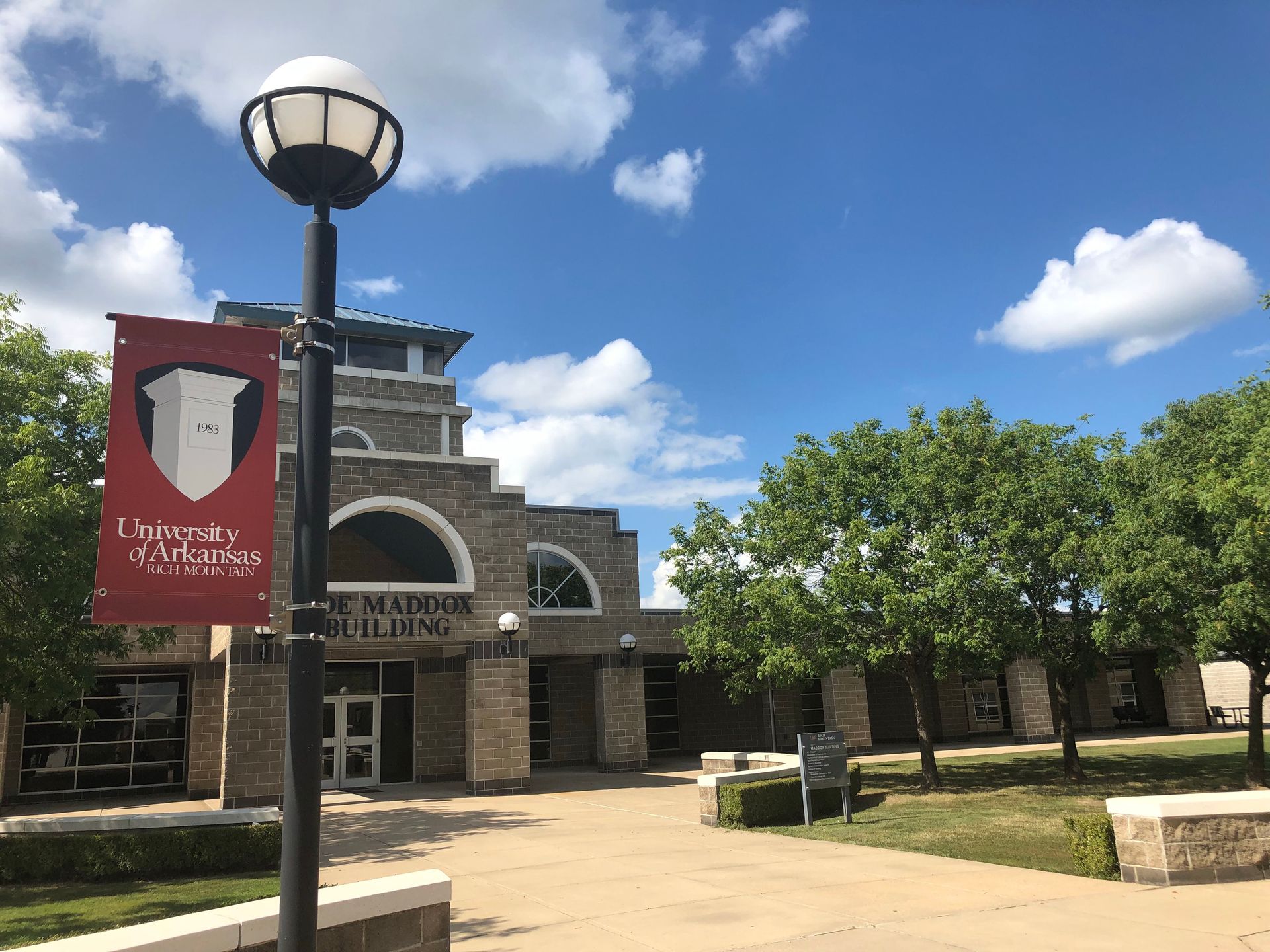 Campus building entrance with stone facade, lamp post banner, trees, and bright blue sky