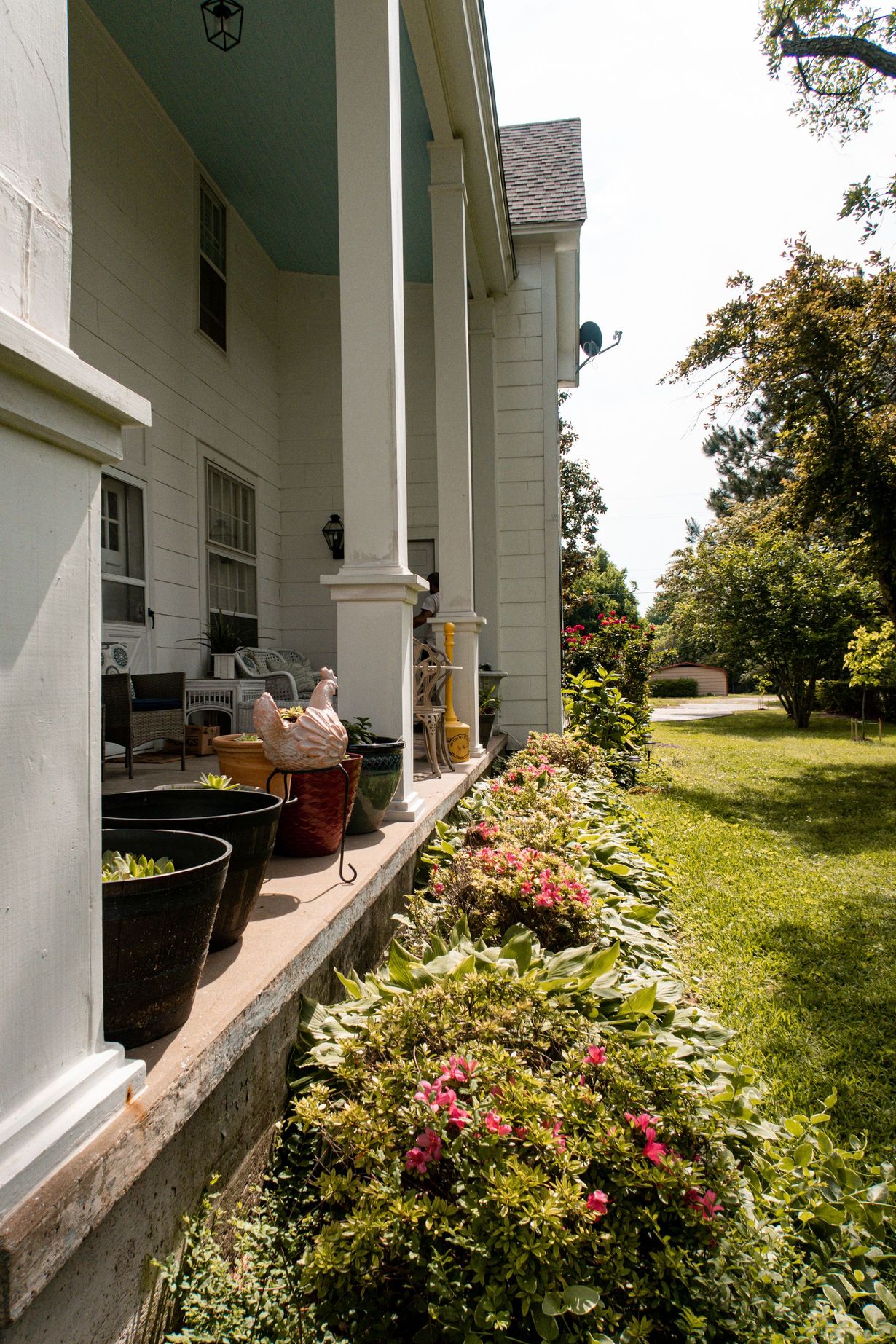 White house porch with potted plants and a flower bed beside a sunlit lawn and trees.