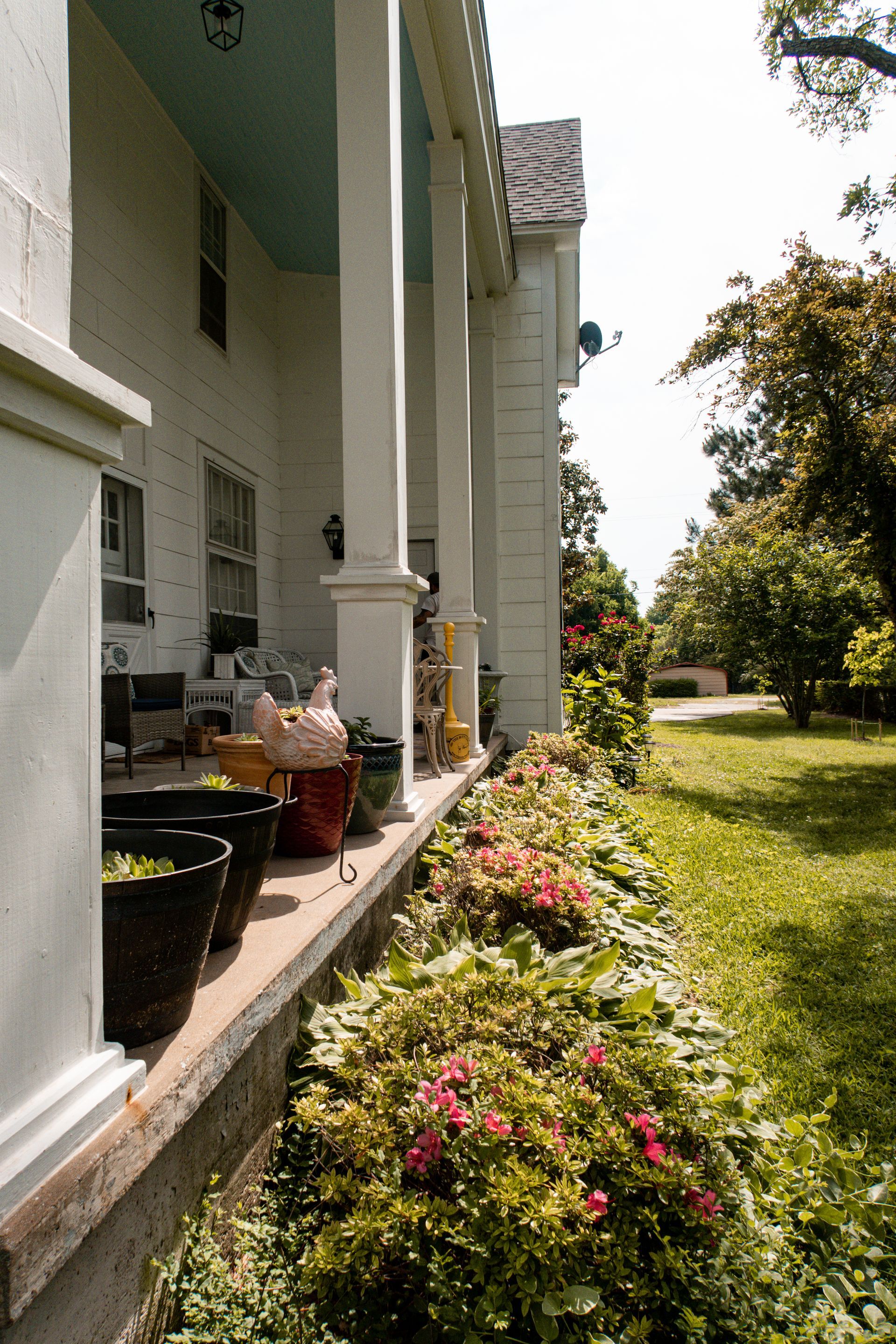 White porch with potted plants and pink flowers beside a sunny green lawn