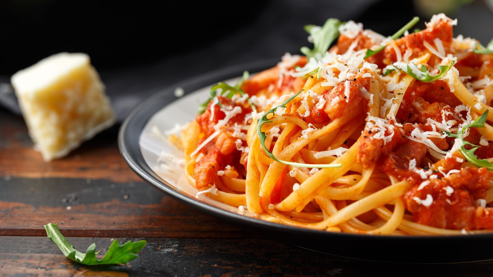 Plate of spaghetti with tomato sauce and grated cheese, with a wedge of Parmesan on a dark table.