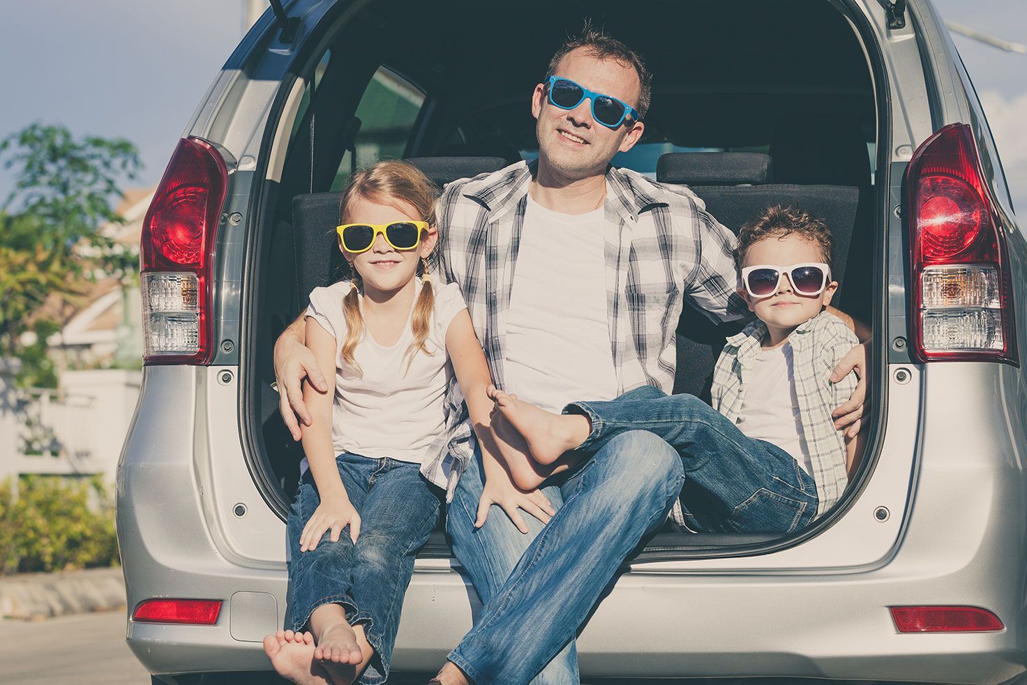 Three people sitting in the open trunk of a silver SUV, wearing sunglasses outdoors.