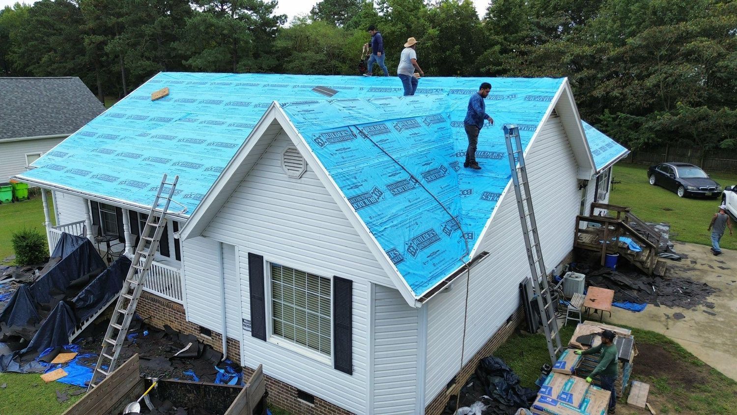 Workers installing bright blue roofing on a white house with ladders and building materials around it