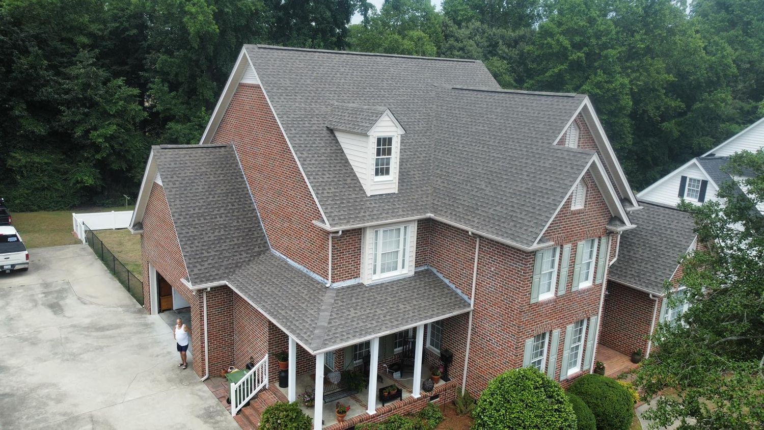 Aerial view of a brick suburban house with gray roof, front porch, driveway, and trees surrounding it.