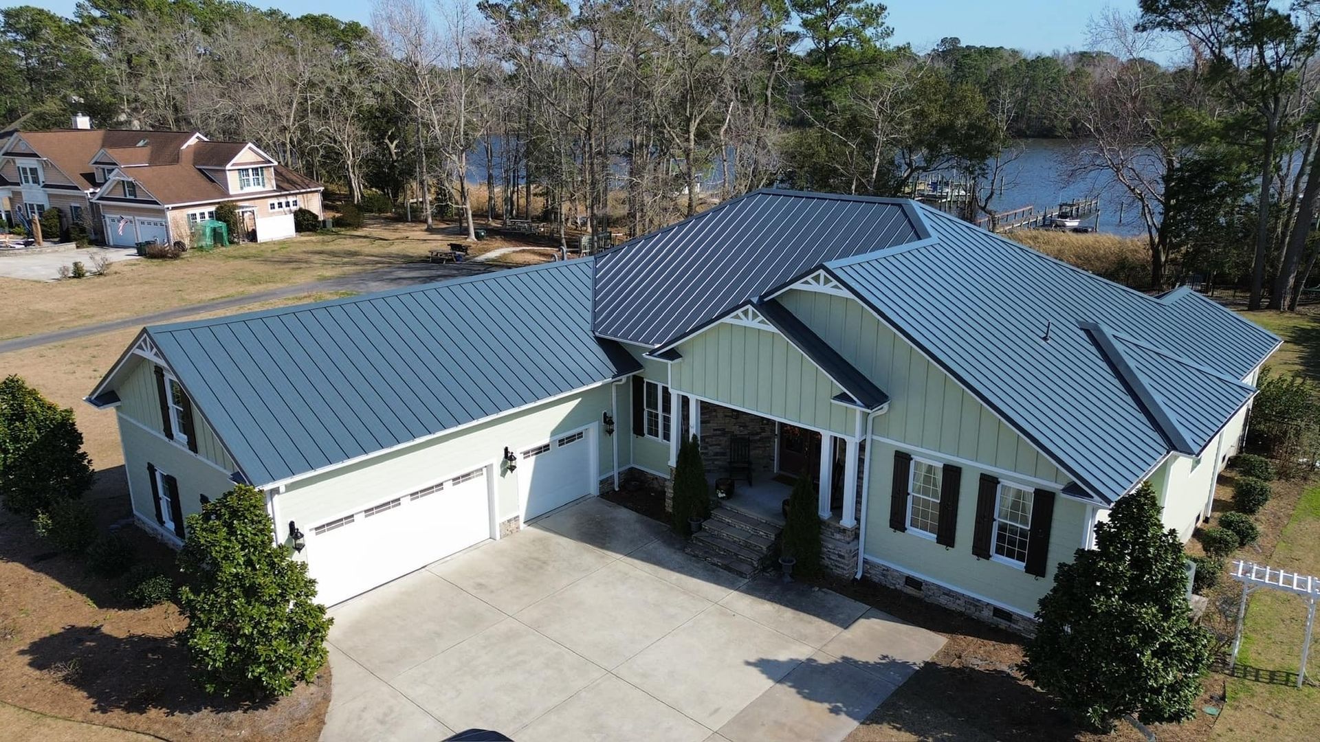 Aerial view of a gray-roofed house with a white garage beside a lake and trees