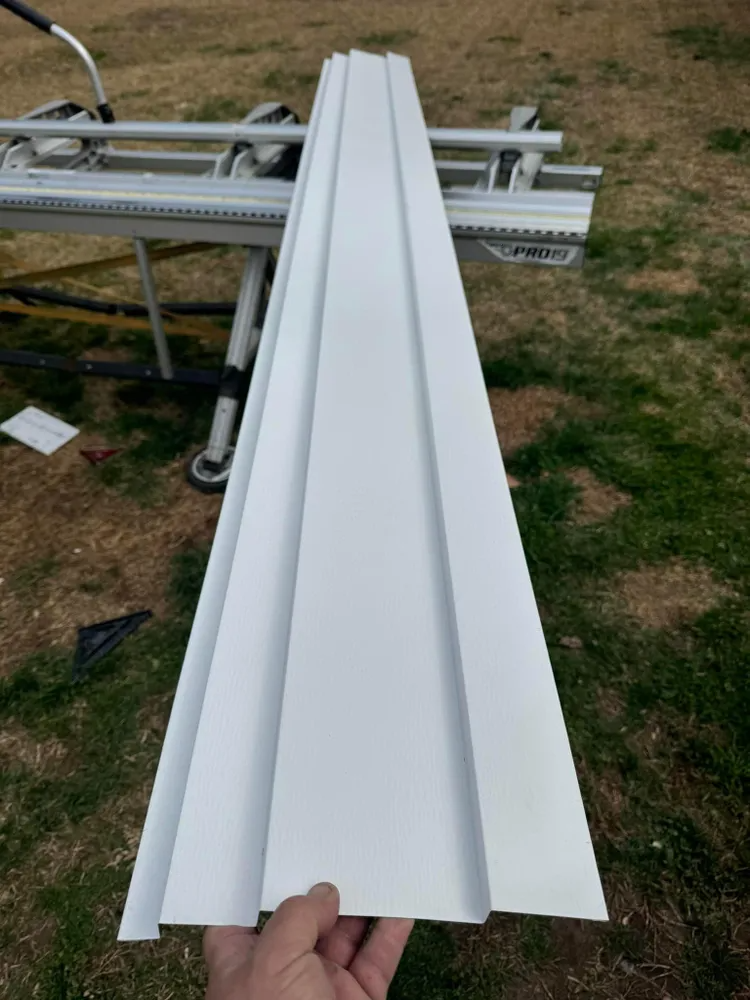 White metal siding panel being held in front of stacked materials outdoors on grass