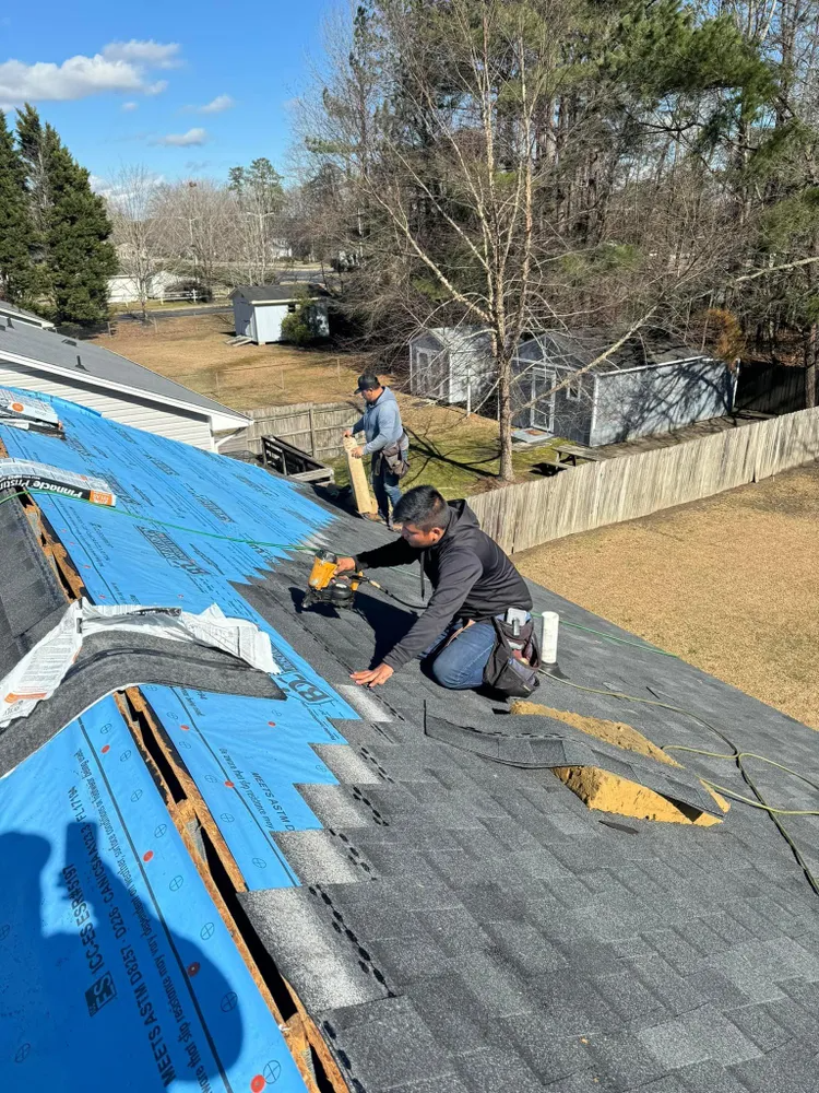 Two workers installing gray shingles on a roof with blue underlayment exposed.