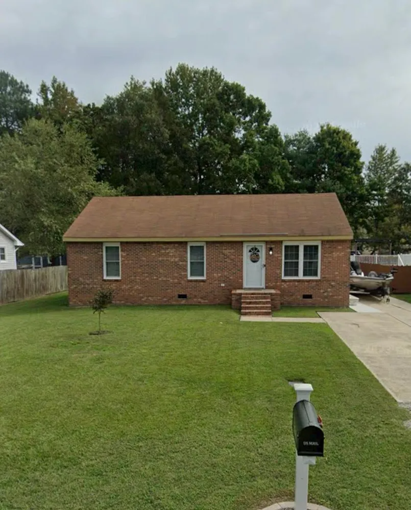 Small brick house with a front lawn, driveway, and mailbox under cloudy skies.