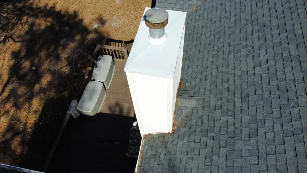 Roof vent pipe on a shingled roof beside a chimney and tree shadow