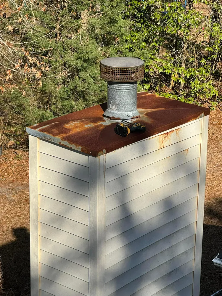 White outdoor cabinet with a rusty metal top and vented chimney, set beside bushes in sunlight
