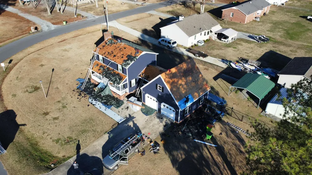 Aerial view of a damaged blue house with a torn roof and debris in a rural neighborhood.