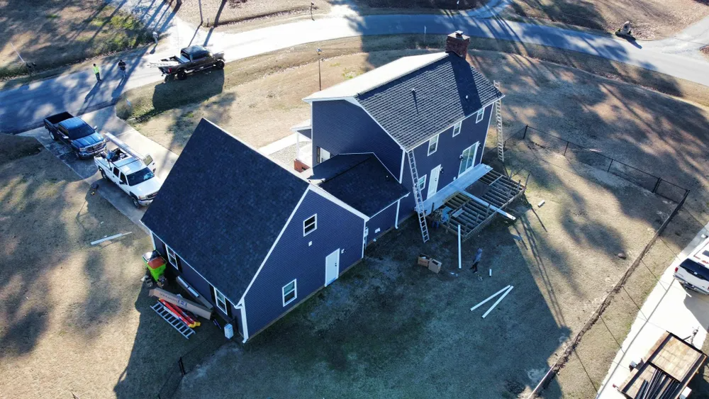Aerial view of a dark blue house with a small deck, yard, and parked vehicles nearby.