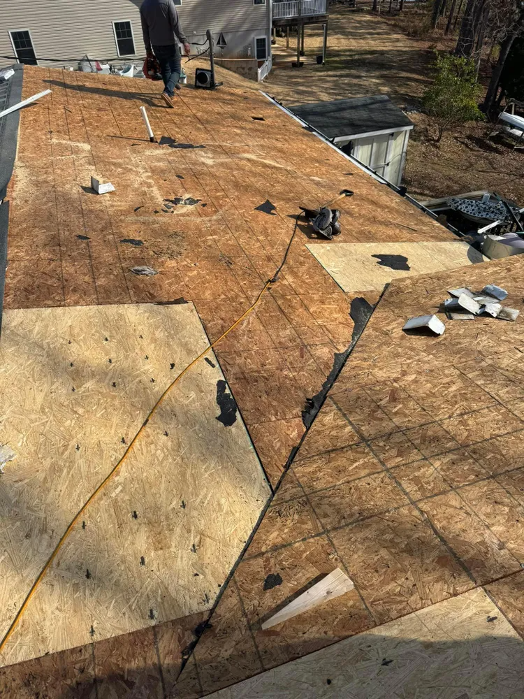 Damaged rooftop with patchy plywood, scattered debris, and a few workers in the distance